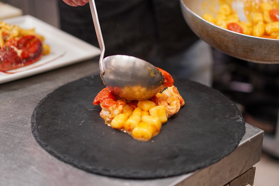 In cucina: un cuoco versa col mestolo il sugo di pomodorino e gamberi sugli gnocchi appoggiati su un piatto di ardesia
