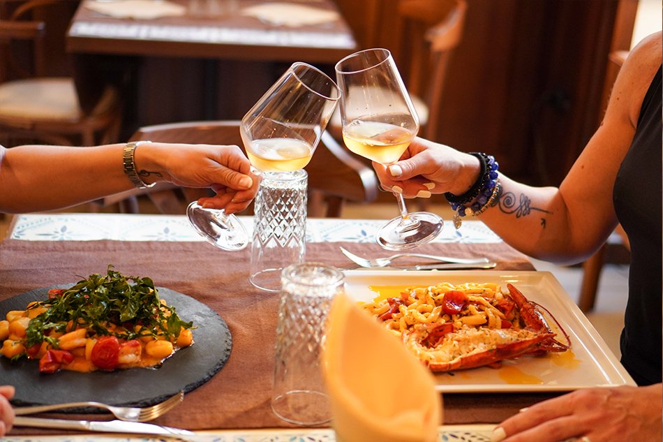 Due persone brindano con il vino bianco sopra il tavolo: sull’ardesia gli gnocchi, sul piatto bianco la chitarrina all’astice